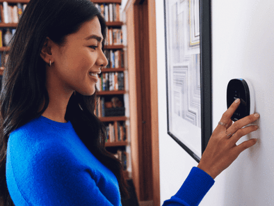 A woman wearing a blue shirt adjusts a thermostat on the living room wall.